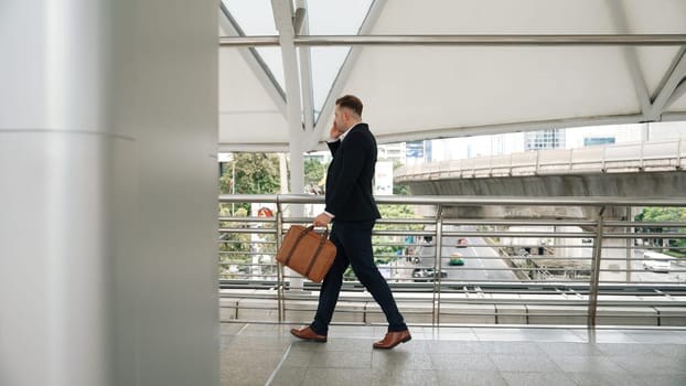 Smart caucasian businessman calling his colleague to plan financial strategy while walking to workplace. Side view of manager using mobile phone to communicate with marketing team. Lifestyle. Urbane.