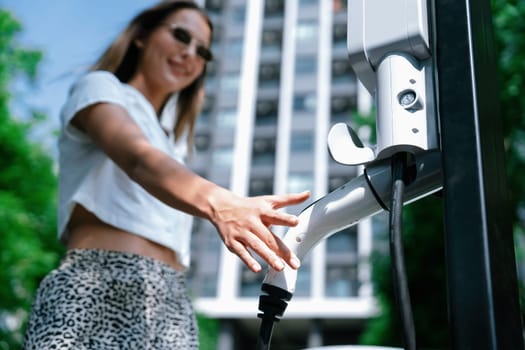 Young woman travel with EV electric car charging in green sustainable city outdoor garden in summer. Urban sustainability lifestyle by green clean rechargeable energy of electric BEV vehicle innards