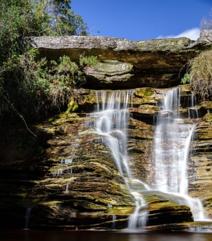 Impressive view of a silky waterfall descending jagged rocks, featuring a large stone with a hat-like hole amid lush greenery.