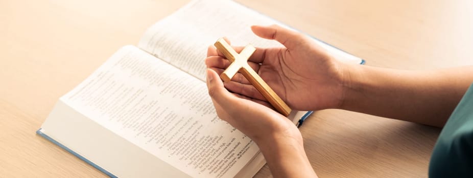 Female god believer holding wooden cross on opened holy bible book at light wooden church table. Top view. Concept of hope, religion, faith, christianity and god blessing. Warm background. Burgeoning.