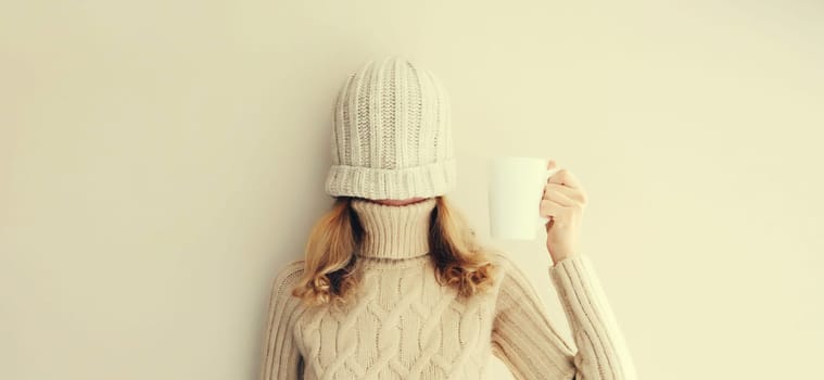 Cheerful funny woman having fun, pulling winter hat over her face warm up holding cup of hot coffee wearing warm soft knitted clothes, sweater on beige studio background