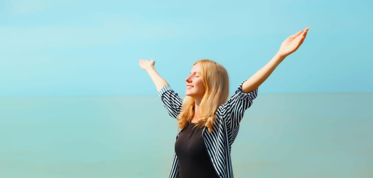 Summer vacation, happy young woman raising her hands up on the beach on sea coast and blue sky background