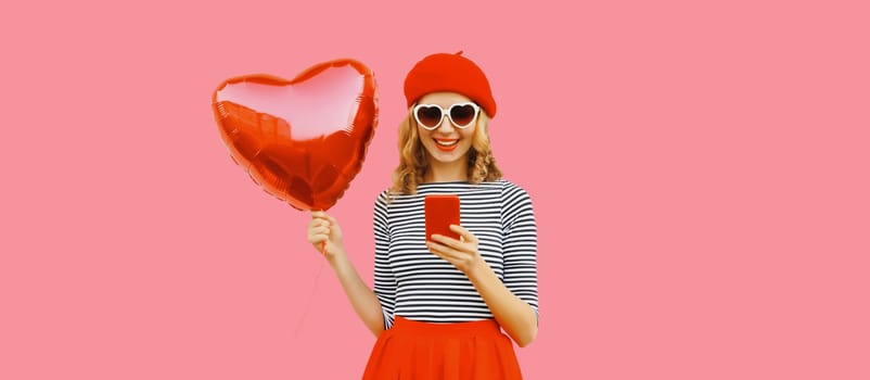 pretty happy smiling young woman with mobile phone and red heart shaped balloon wearing french beret, sunglasses on pink studio background