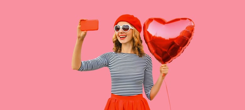 happy smiling young woman taking selfie with mobile phone holding red heart shaped balloon wearing french beret, sunglasses on pink studio background