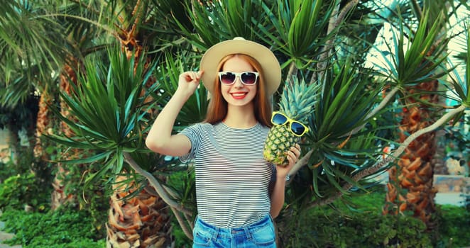 Summer portrait of happy smiling young woman with pineapple fruits wearing sunglasses, straw hat posing on palm tree background