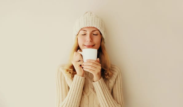 Winter portrait of happy satisfied young woman enjoying cup of hot tasty coffee or tea in warm knitted sweater and hat in early morning at home
