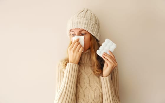 Sick woman sneezing blow nose using tissue and holding pills wearing warm soft knitted clothes, hat and sweater on beige studio background