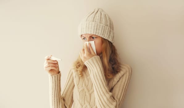 Sick woman sneezing blow nose using tissue and measuring checking body temperature looking at thermometer wearing warm soft knitted clothes, hat and sweater