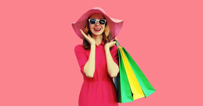 Portrait of beautiful happy smiling young woman model posing with colorful shopping bags in summer straw hat, dress on pink studio background