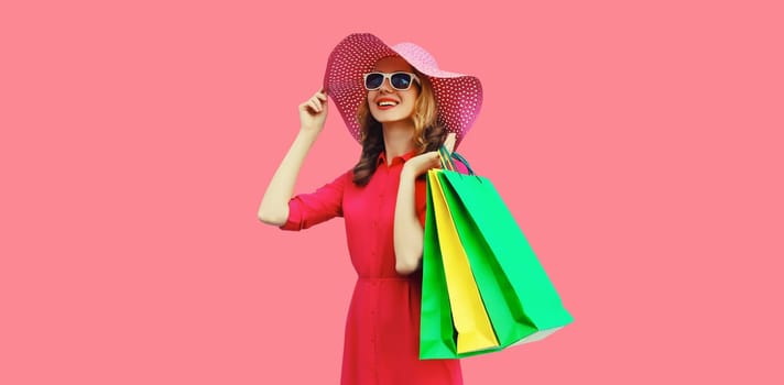 Portrait of beautiful happy smiling young woman model posing with colorful shopping bags in summer straw hat, dress on pink studio background