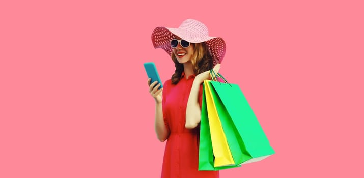 Beautiful happy young woman looking at mobile phone with colorful shopping bag in summer straw hat on pink studio background