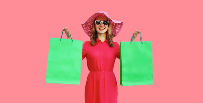 Portrait of beautiful happy smiling young woman model posing with colorful shopping bags in summer straw hat, dress on pink studio background