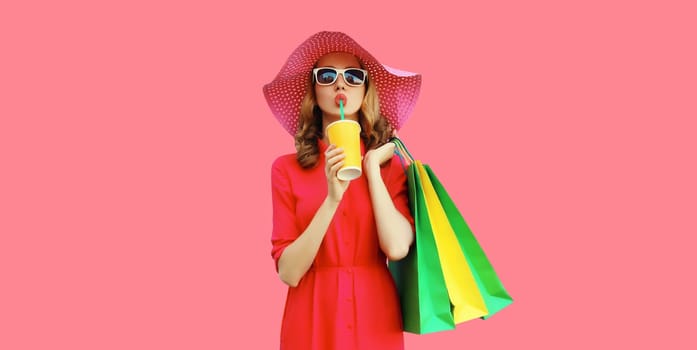 Portrait of beautiful relaxed young woman model with colorful shopping bags drinking juice in summer straw hat, dress on pink studio background