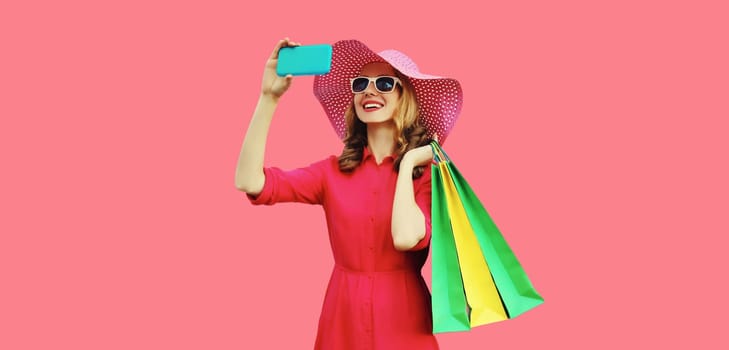 Beautiful happy smiling young woman model taking selfie with mobile phone holding colorful shopping bags in summer straw hat, dress on pink background