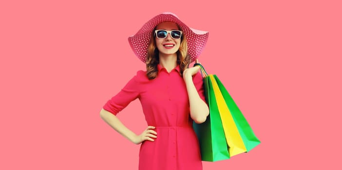 Portrait of beautiful happy smiling young woman model posing with colorful shopping bags in summer straw hat, dress on pink studio background