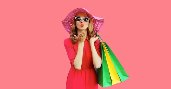 Portrait of beautiful happy young woman model blowing kiss posing with colorful shopping bags in summer straw hat, dress on pink studio background