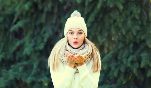 Winter portrait of happy cheerful blonde young woman blowing snowflakes from hands in hat, sweater in forest on christmas tree background