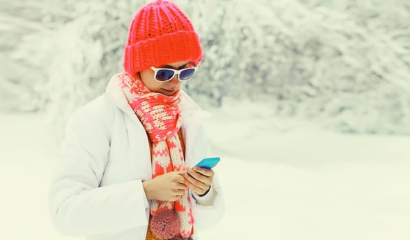 Winter portrait of happy young woman with mobile phone in knitted hat, scarf and jacket in snowy park