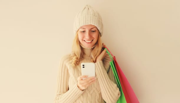 Happy smiling young woman looking at mobile phone with shopping bag in winter knitted hat and warm sweater