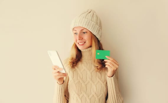 Happy young woman shopping online or ordering something looking at smartphone holds plastic credit bank card in winter knitted hat, sweater on beige background
