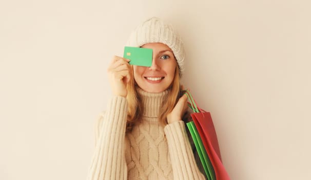 Portrait of happy smiling young woman holding plastic credit bank card with shopping bags in winter knitted hat and sweater on beige studio background