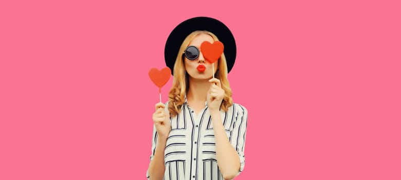 Portrait of happy beautiful young woman blowing her lips sends kiss with sweet red heart shaped lollipop on stick in black round hat on pink studio background