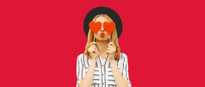 Portrait of happy beautiful young woman blowing her lips sends kiss with sweet red heart shaped lollipop on stick in black round hat on pink studio background