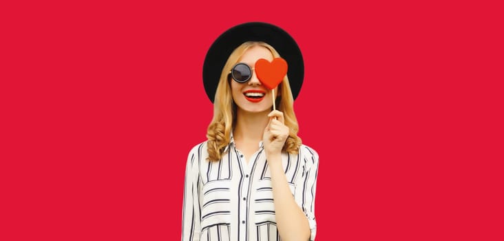 Portrait of happy cheerful young woman with sweet red heart shaped lollipop on stick in black round hat on pink studio background