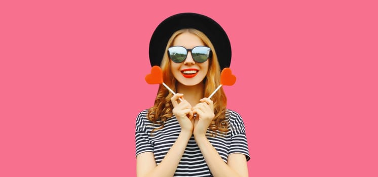 Portrait of happy cheerful young woman with sweet red heart shaped lollipop on stick in black round hat on pink studio background