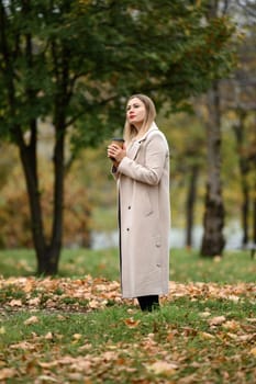 A young girl in a long autumn coat drinks coffee in an autumn park, portrait photo of a girl in the park.