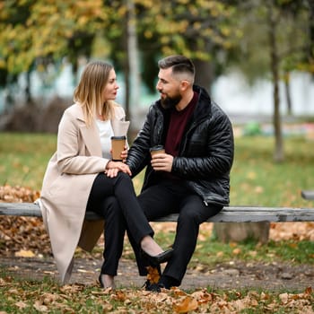 A couple, a man and a woman, are sitting in the park on a bench with coffee in their hands, a meeting in the park.