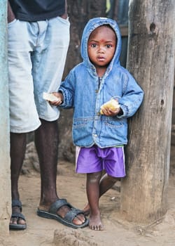 Ranohira, Madagascar - April 29, 2019: Unknown Malagasy boy, in blue denim jacket with hood and shorts, barefoot, standing next to his father legs, holding bread in hands. Locals are poor but cheerful
