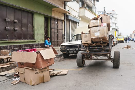 Antananarivo, Madagascar - April 24, 2019: Simple cart with empty carton boxes and wooden boards (to setup stall for morning market) pulled at main street. Unknown Malagasy man sits on floor at back
