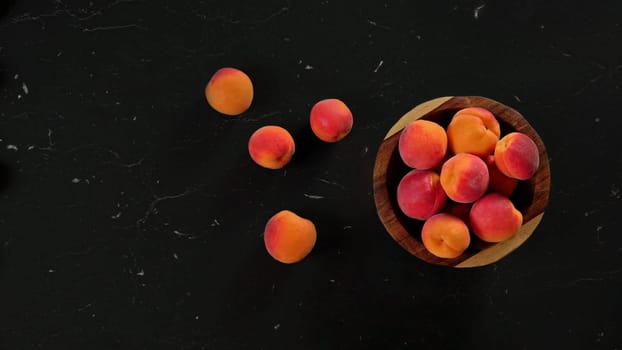 Ripe apricots in small wooden bowl, some scattered on black marble like desk next to it. View from above, space for text left
