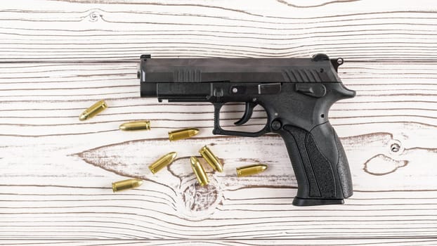 Simple black modern pistol gun with few yellow brass bullets on white wooden board, view from above
