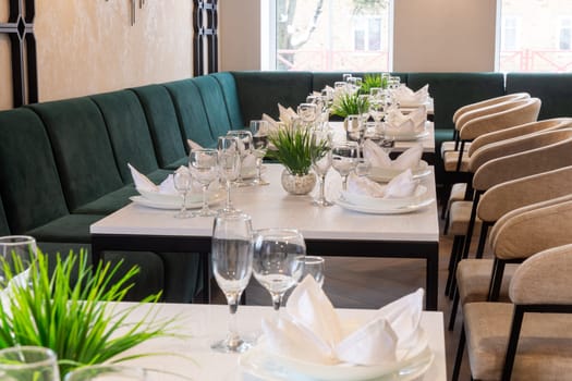 Modern interior of urban restaurant or cafe with dining places. Soft green sofas and beige chairs around tables served with glasses, plates and napkins.