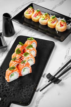 Two types of sushi rolls served with soy sauce on black plates against marble background. In focus classic Philadelphia rolls with cream cheese, cucumbers and salmon topped with crispy rice balls