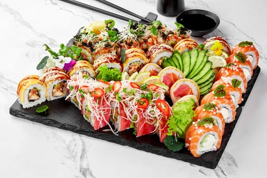 Diverse selection of sushi with eel, salmon and tuna beautifully garnished with edible flowers, greens, cucumber, shredded daikon and chili, artistically arranged on slate board, served with soy sauce