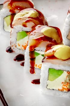 Closeup of sushi rolls filled with cream cheese, tobiko and avocado topped with sea scallop and drizzled with spicy mayo and savory unagi sauce, served on plate. Japanese cuisine