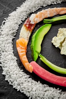 Closeup of colorful half circle of slices of fresh raw salmon, tuna, eel, shrimps, ripe avocado, cucumber, white rice and Japanese mayonnaise on black textured background. Ingredients for making sushi