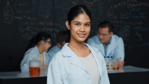 Girl looking at camera with arm folded while people doing experiment at laboratory. Cute student standing blackboard with chemical theory with blurring background at STEM science class. Edification.