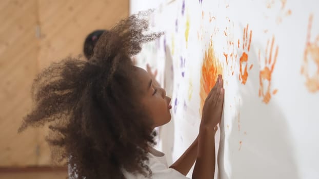 African girl paint the wall with colorful stained hands with friend. Group of multicultural learner standing in front of wall while using hand to create artwork. Creative activity concept. Erudition.