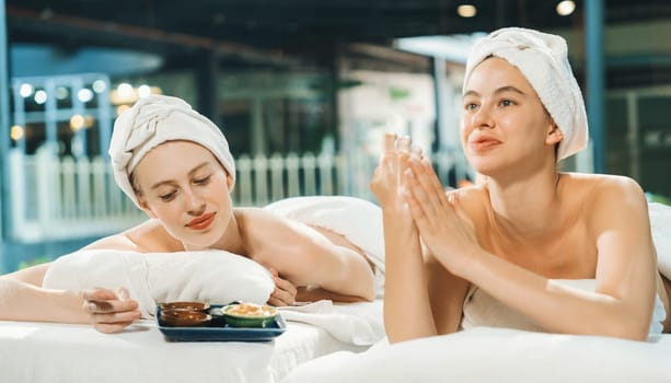 Couple of beautiful young girls lie on spa bed during interested in homemade beauty facial mask. Attractive woman touching herbal facial mask. Surrounded with nature environment. Tranquility.