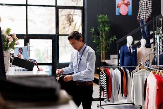 Asian store employee arranging clothes, putting fashionable merchandise on hangers, working at modern boutique visual. Stylish man checking formal wear items in shopping centre. Fashion concept