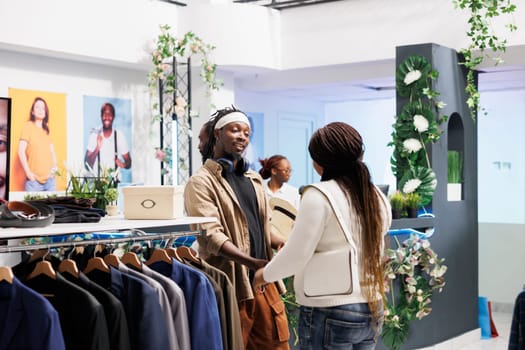 Woman buying trendy accessories and asking boyfriend for advice in shopping center. African american couple customers choosing stylish hat and clothes in mall fashion boutique