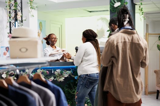African american customers waiting in line at cash register to complete purchase in clothing store. Cashier taking apparel to scan and process payment at shopping mall checkout counter