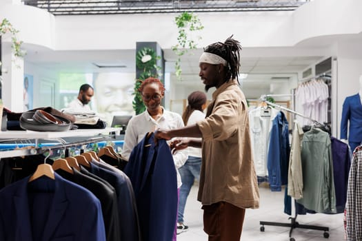 African american buyer and seller discussing fashion trends while choosing jacket in clothing store. Shopping mall customer and assistant browsing through rack with hanging blazers