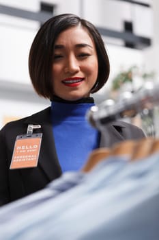 Fashion boutique smiling asian woman seller with badge examining formal shirts on display rack. Department shopping center clothing store assistant managing garment inventory