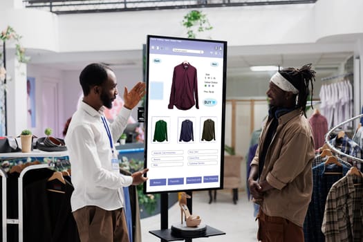 Young worker showing items on touch screen monitor kiosk service to client in fashion boutique at mall. Store assistant advising customer to select trendy clothing line merchandise.