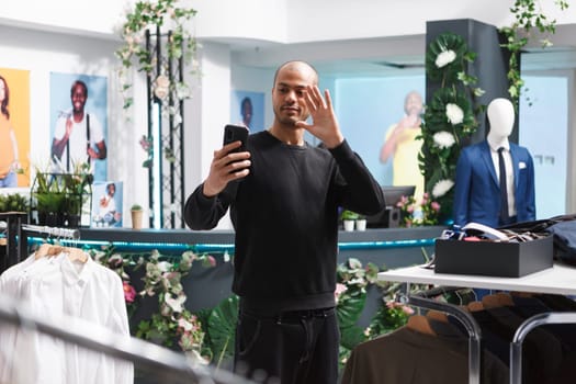 Arab man customer in clothing store holding smartphone, looking at front camera and waving with hand while shopping for apparel. Influencer greeting followers while streaming in boutique